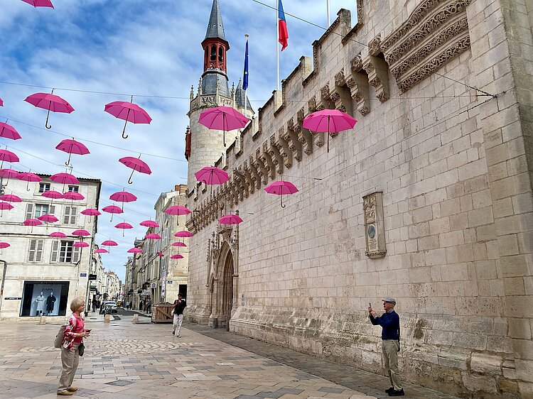 Straßenszene mit historischen Gebäuden und pinken Regenschirmen, die über einer Altstadt aufgehängt sind.