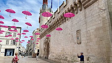 Straßenszene mit historischen Gebäuden und pinken Regenschirmen, die über einer Altstadt aufgehängt sind.