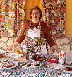 Kochkurs in Sizilien: festlich gedeckter Tisch mit Süßspeisen, ältere Frau in traditioneller Kleidung vor gemusterter Wand.