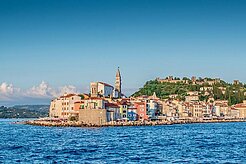 Küstenstadt Piran mit Kirchturm, bunte Häuser am Meer unter blauem Himmel.