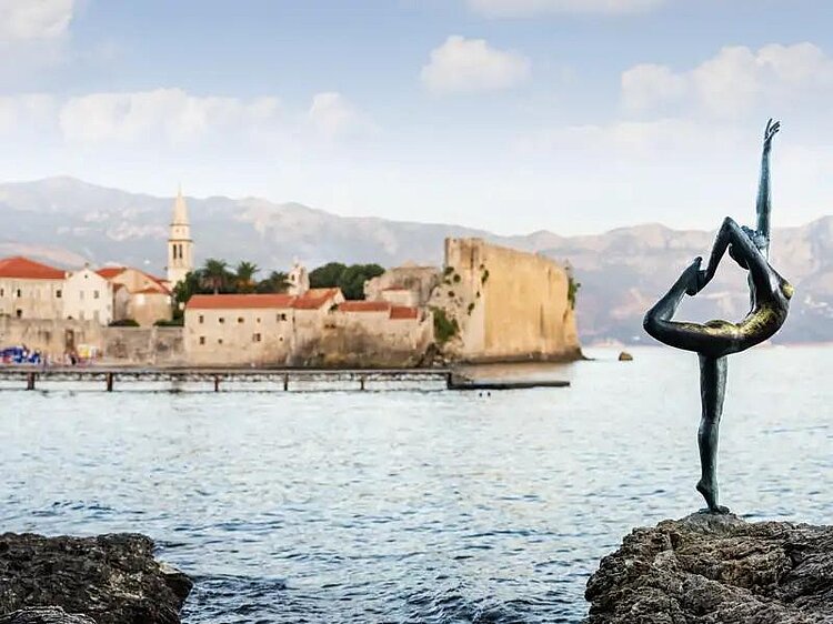 Bronzene Ballerina‑Skulptur am Ufer von Budva, Montenegro, Altstadt im Hintergrund.