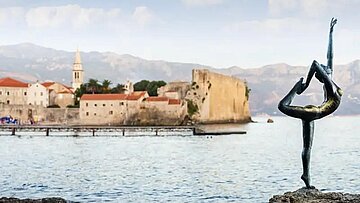 Bronzene Ballerina‑Skulptur am Ufer von Budva, Montenegro, Altstadt im Hintergrund.